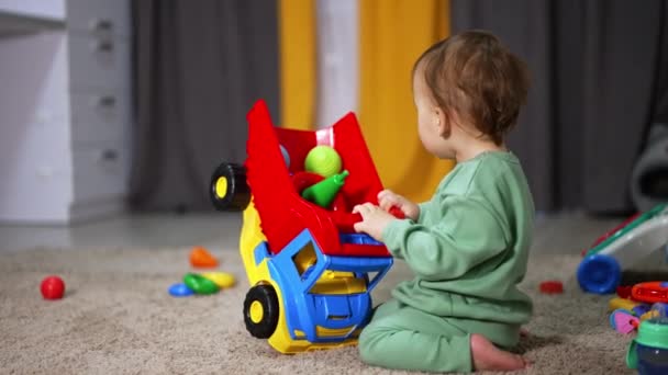 柔らかいカーペットの上に明るいローリーで遊ぶ甘い子供 トッドラーは車のバスケットからおもちゃを取り出すために車を回します ブラーレッド バックドロップ — ストック動画