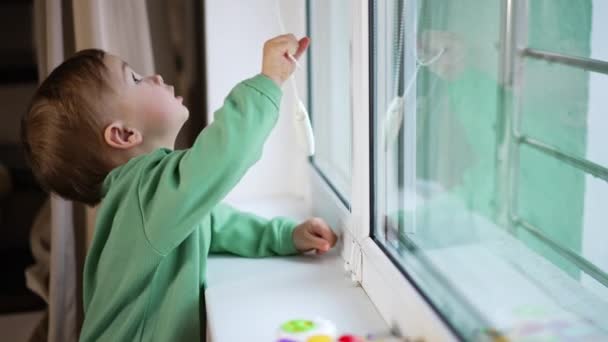 明るい緑色の服を着た小さな赤ん坊が窓のそばに立っている かわいい子供は嫉妬深い窓のカーテンで幸せに遊んでいます — ストック動画