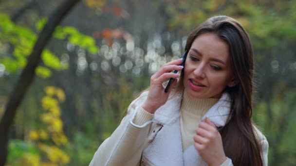 笑顔の女性が屋外で電話で話しています 魅力的なブルネットは秋の公園で彼女の携帯電話にセルフィーを取ります ブラーレッド バックドロップ — ストック動画