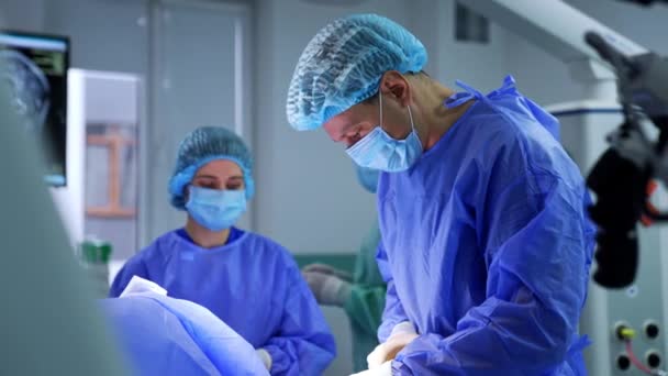 男性の外科医は 彼にショットを与える操作テーブルの上に患者を曲げました 手術のための材料を準備する医療スタッフ — ストック動画