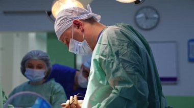 Kafkasyalı doktor elektrikli aletle hastanın üzerine eğildi ve hemşireye uzattı. Anestezi uzmanı arka planda operasyonu izliyor..