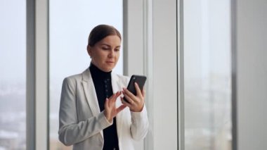 İş kadını pencerede duruyor ve telefon görüşmesi yapıyor. Beyaz ceketli kadın telefonuna bakar, eliyle konuşur ve el kol hareketi yapar..