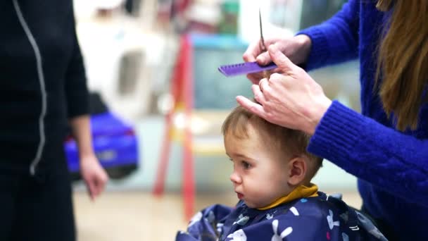 サロンで1歳の幼児のための髪の切断 青いセーターの長髪のバーバーは 帽子をかぶった小さな男の子の髪をカット — ストック動画