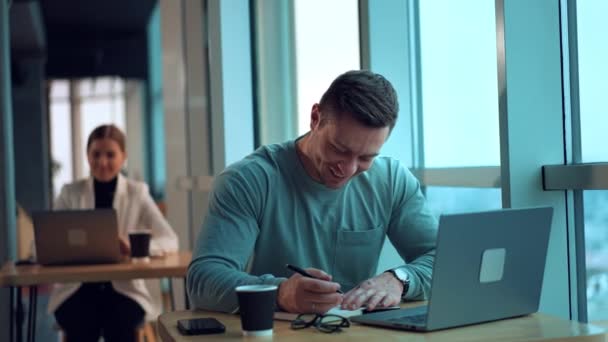 ポジティブな笑顔の男は紙のノートにノートを取る 男性のオフィスの従業員はノートパソコンを見て 誰かに話しかける バックドロップでコンピュータで働く女性の同僚 — ストック動画