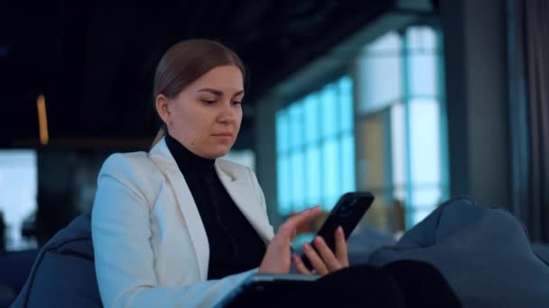 白いジャケットを着ているビジネスレディは 彼女の携帯電話に焦点を当てた快適なアームチェアに座っています スマートフォンを使って働く白人女性 クローズアップ ブラーレッド バックドロップ — ストック動画