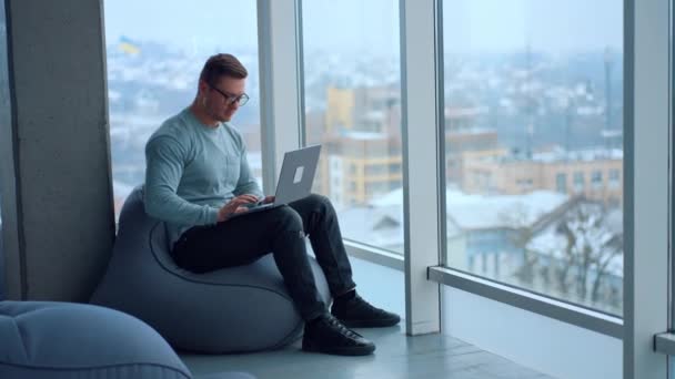 男自由职业者舒适地坐在大玻璃窗前的豆袋椅上 在一个视野开阔的房间里工作的人 — 图库视频影像