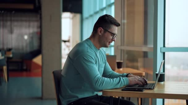 カフェに座っているノートパソコンで働くフリーランスの男性 リラックスした雰囲気の中でコンピュータにタイピングするメガネの男性 ブラーレッド バックドロップ — ストック動画