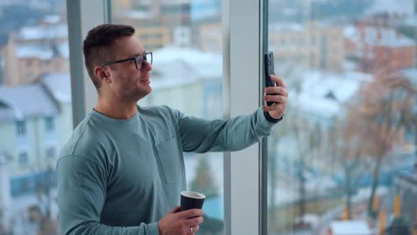 窓に立っている手の紙のコップと眼鏡をかけた男 スマートフォンでビデオチャットを持っている白人男性 — ストック動画