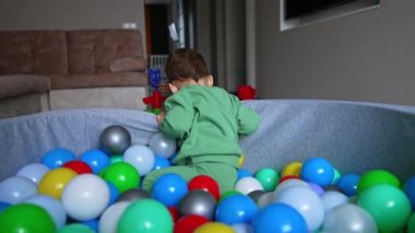 Toplarla dolu yumuşak havuzun içinde oturan neşeli küçük çocuk. Tatlı çocuk yeşil topu almak için havuzun dışına uzanıyor..