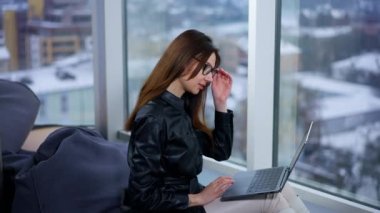 Laptop 'ta çalışan koyu renk saçlı, kendine güvenen bir bayan. Kadın gözlüklerini çıkarıp pencereye bakıyor..