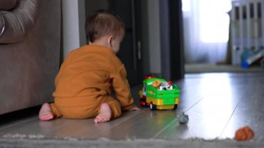 Oyuncak araba yerde hareket ediyor ve çıplak ayaklı bebek onu takip ediyor. Çocuk dizlerinin üstüne çöker ve oyuncağın düğmesine basar. Arka görünüm.