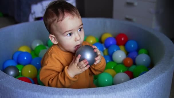 可爱的一岁婴儿和玩具一起坐在干水池里 孩子拿着一个球试图咬它 靠近点 — 图库视频影像