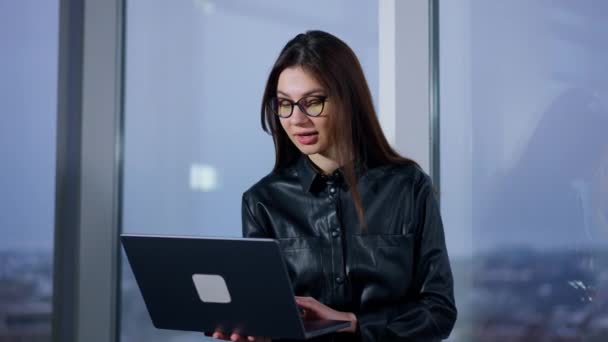 戴着眼镜的迷人的女商人看着站在窗前的笔记本电脑 在网上聊天的女士对着摄像机笑 — 图库视频影像