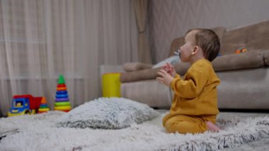 Bir yaşında bir çocuk dizlerinin üstüne çöküp yere oturuyor. Bebek çocuk elinde ve ağzında tuttuğu emziği çiğniyor..