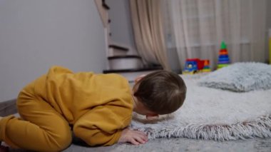 Beyaz bir çocuk halıya uzanıp sırtüstü yatıyor. Çocuk kendini ayak parmaklarından tutarak bacaklarını kaldırır. Kapat..