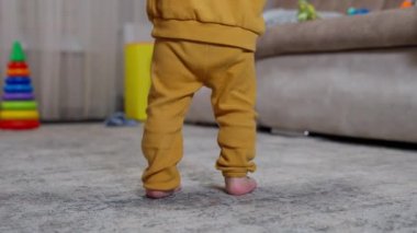 Turuncu takım elbiseli çocuk içeride yürümeyi öğreniyor. Bebek, annesi tarafından desteklenerek ayaklarını kaldırıyor..