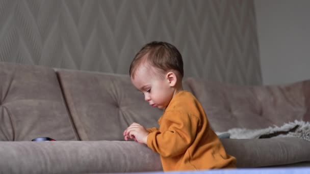 大きなソファーの近くに立っているオレンジ色のスポーツスーツの白人少年 子供は彼の手の白い箱に注意深く見える — ストック動画