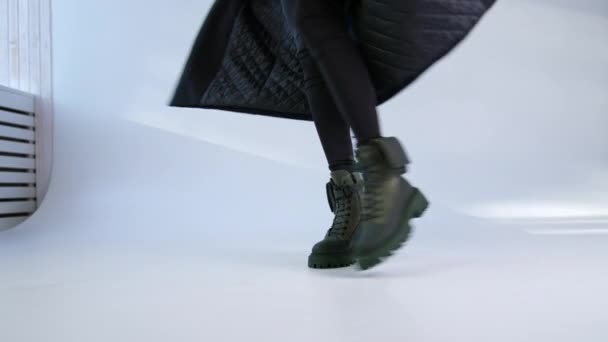 身穿黑色长外套的女人穿着时髦的战斗靴走在工作室旁边 两边都是小包 白色背景 — 图库视频影像