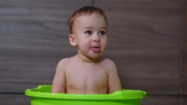 面白いかわいい赤ん坊の男の子は舌を示す浴槽に座っています 子供はそこに手を伸ばして見上げた クローズアップ — ストック動画