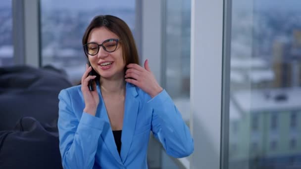 一位成功的有弹性的商务女士坐在办公室里 通过电话交谈 穿着蓝色夹克和眼镜的布鲁内特女人看着窗户 一边聊天一边摆姿势 — 图库视频影像