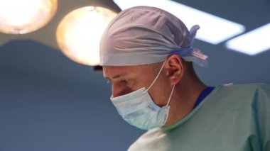 Maskeli ve şapka takan yetişkin bir cerrah. Kapatın. Ameliyat odasındaki lambalar bulanık arkaplanda yanıyor..