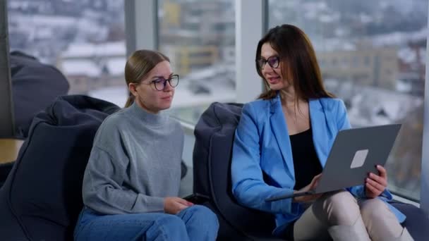 青いジャケットの女性が膝の上にラップトップを持っていて スクリーンに何かを同僚に見せている オフィスでの女性社員のコミュニケーション ブラーレッド バックドロップ — ストック動画