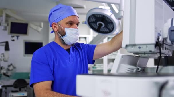 マスクとブルーユニフォームの男性医師は 肺換気機械のスクリーンを回転させます 現代の手術の準備をする医学 — ストック動画