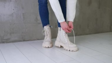 Kot pantolon giyen, beyaz kazak giyen, dantellerini beyaz botlara bağlayan tanınmamış bir kadın. Model gri duvarın yanında duruyor..