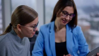 Modern ofiste oturan kadın çalışanlar ekrana odaklandı. Mavi ceketli kadın dizüstü bilgisayarı işaret eden bir şeyi açıklıyor ve diğeri onu dinliyor..