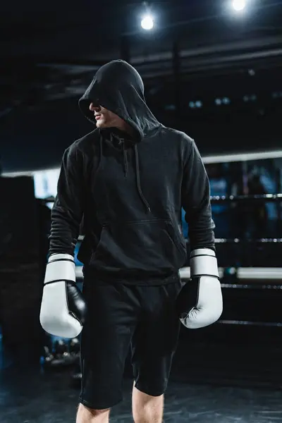 Young man standing in the boxing gym with hood on his head and boxing gloves on his hands.