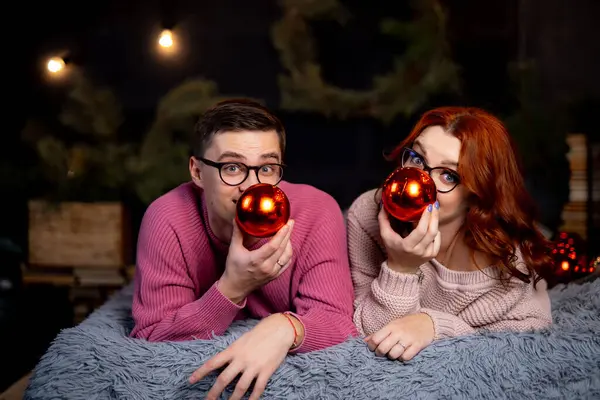 Young couple is lying on blanket and holding Christmas balls in their hands.
