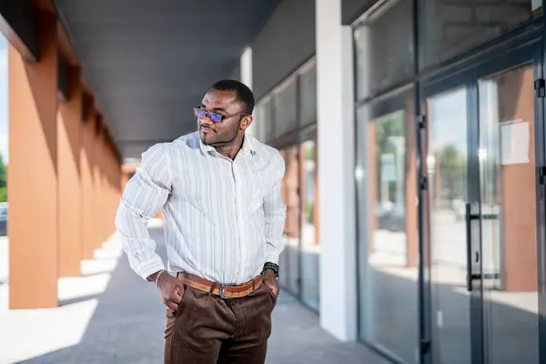 Stylish African American man in white shirt and brown pants stands on the street near the building and looks away. African American guy is resting after long day., Stylish African American man in white shirt and brown pants stands on the street near