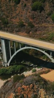 Köprü ve arabalar hareket halinde. Küçük derenin üstündeki dağlarda kemerli köprü. Hava görüntüsü. Dikey video.