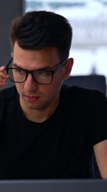 Genç beyaz adam bilgisayar başında çalışmak için gözlük takıyor. Freelancer geniş odada otururken dizüstü bilgisayarında daktilo yazıyor. Arkaplan bulanık. Dikey video.