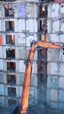 Kazı makinesi eski bir binanın duvarlarını sökmek için yükseğe ulaşır. Beton ve ağır toz parçaları dökülüyor. İnşaat kazıcıları eski bir binanın kalıntılarını temizliyorlar. Dikey video.