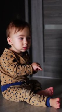 Sevimli küçük erkek bebek içeride yerde oturuyor. Meraklı çocuk kapalı kapıya döner ve ona bakar. Dikey video.