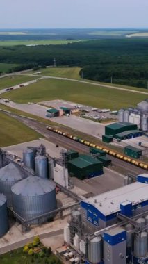 Tahıl depolama ve işleme için modern teknoloji fabrikası. İyi korunan sanayi bölgeleri kargo trenleriyle demiryollarına bölünmüş durumda. Güzel doğa manzarası arka planı. Dikey video.