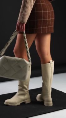 Kısa etek ve süveter giyen kız açık renkli çizmeler ve aynı renk çanta gösteriyor. Stüdyoda ayakkabı ve aksesuar gösterisi. Dikey video.