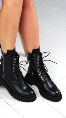 Taburede oturan bej ceketli kadın bağcıklı siyah çizme gösteriyor. Güzel şık deri çizmeler sunumu. Beyaz zemin. Dikey video.