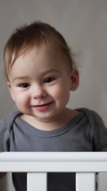 Beyaz karyolada duran gri tişörtlü güzel çocuk. Gülümseyen bebek elini tatlı tatlı sallıyor. Kapatın. Dikey video.