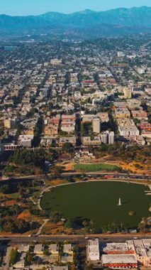 Şehir panoramasının ortasında göleti olan sevimli bir park. Kaliforniya dağlarının arka planında yoğun bir şekilde inşa edilmiş Los Angeles şehir manzarası. Dikey video.