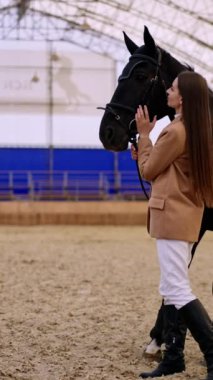 Manege 'de güzel siyah bir atı olan ata binen kadın. Bir kadın atı dizginlerinden tutup hayvanı okşuyor. Dikey video.
