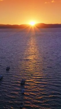 Tahoe Gölü 'nün sularında yüzen çok sayıda tekne var. Parlak güneş arka planda dağların ardında batıyor. Arka planda turuncu gökyüzü. Dikey video.