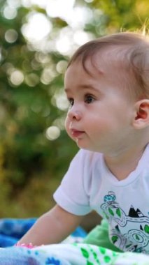 Dört ayak üstünde duran güzel küçük bebek dosdoğru karşıya bakıyor. Beyaz tişörtlü sevimli çocuk bahçede oyuncakla oynuyor. Dikey video.
