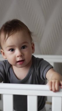 Sevimli çocuk karyolasında ayakta duruyor, tırabzanlardan tutuyor. Güzel çocuk uyandıktan sonra etrafına bakıyor. Arkaplan bulanık. Dikey video.