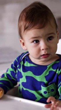 Yeşil lekeli tişörtlü sevimli aktif erkek bebek yemek masasında oturuyor. Küçük çocuk el sallıyor ve sonra bir şeye bakıyor. Kapatın. Dikey video.
