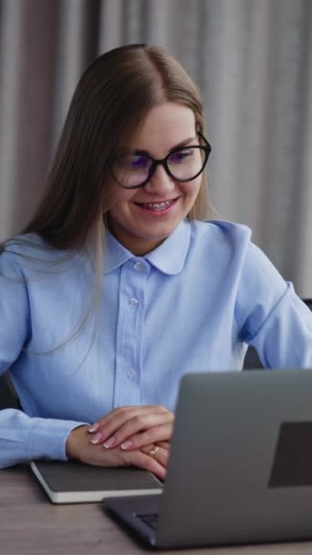 ポジティブな若い女性従業員がコンピュータの情報を調べています レディーはノートパソコンのキーボードに微笑んで入力しています 室内バックドロップ バーティカルビデオ — ストック動画