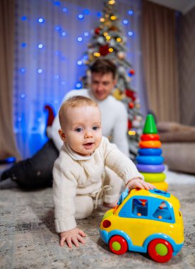 Küçük çocuk, Noel ağacının arka planında yerde oyuncak arabasıyla oynuyor. Baba ve bebek birlikte mutlular..