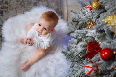 Küçük çocuk Noel ağacının yanındaki kürk halıda oturuyor ve elinde oyuncak tutuyor..