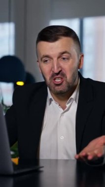 Yakışıklı iş adamı bilgisayarında internette geziniyor ve konuşuyor. Büyük ofisteki bilgisayarda çalışan takım elbiseli bir adam. Arka plan bulanık. Dikey video